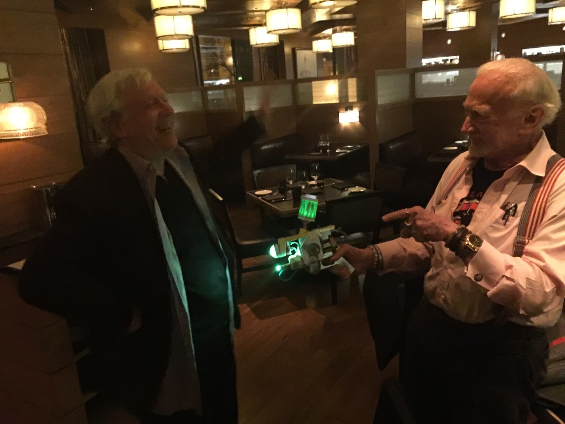 Ralph Benko, at left, laughing as Buzz Aldrin holds up a small illuminated electronic device; the two are standing in a restaurant dining room.