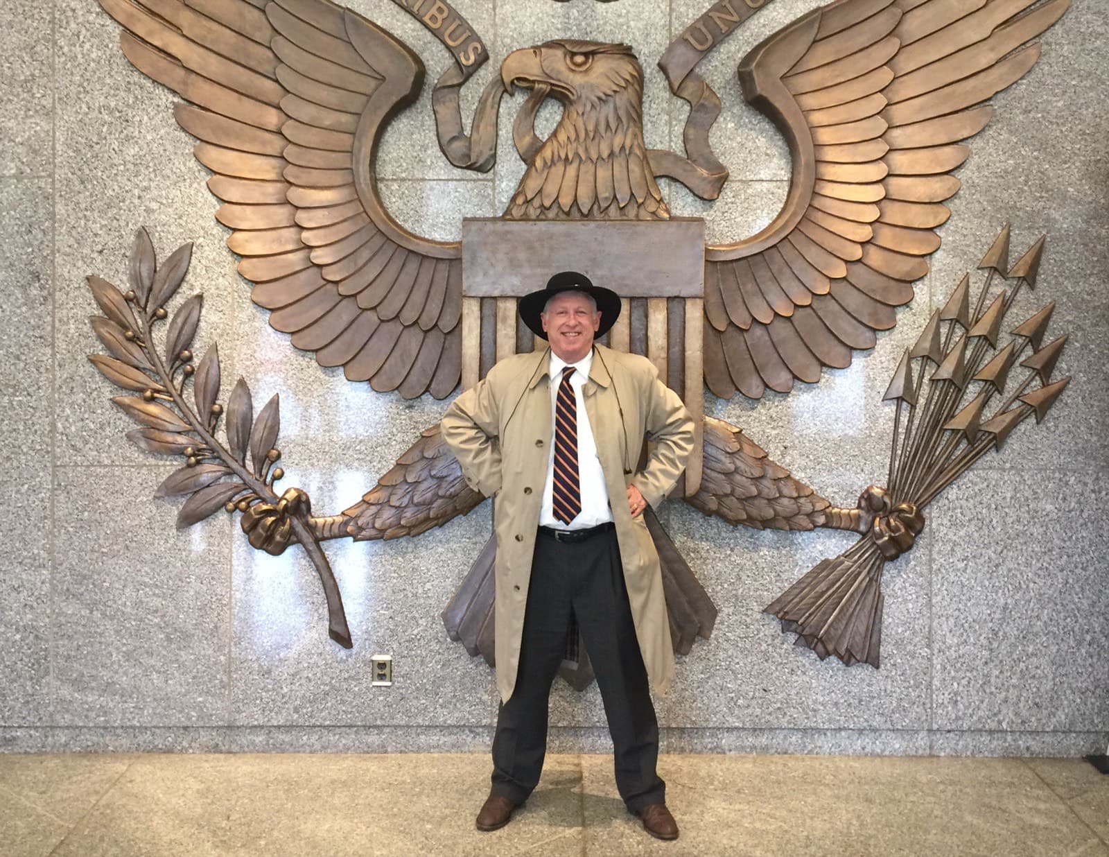 Ralph Benko in a fedora and belted trench coat, hands on his hips, standing before a monumental bronze Great Seal of the United States — the spread-winged eagle holding an olive branch and a bundle of arrows — mounted on a polished granite wall.